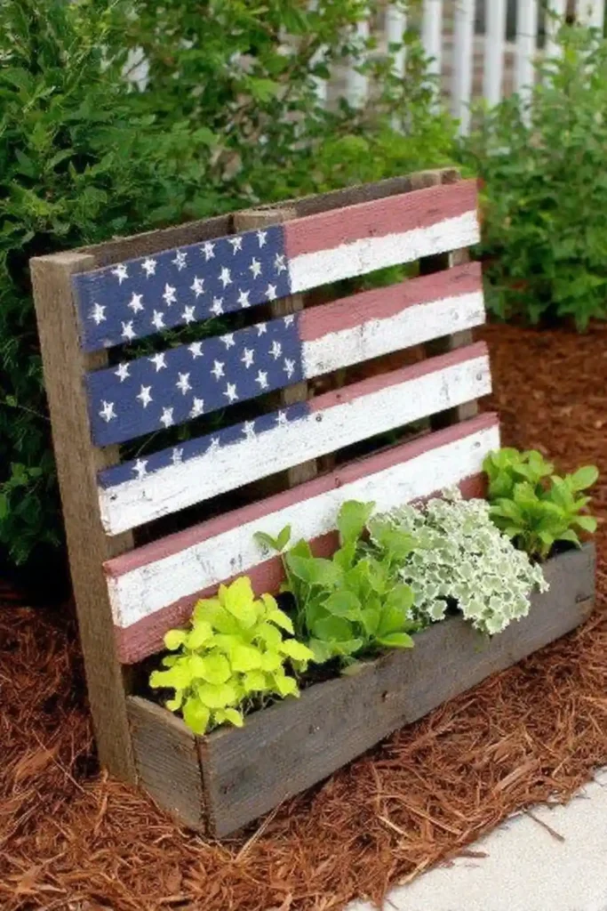 American flag painted pallet planter leaning on mulch with plants behind it