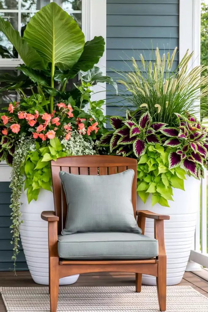 Wooden porch chair with gray cushion between tall planters filled with coral flowers, vines, coleus, and grasses