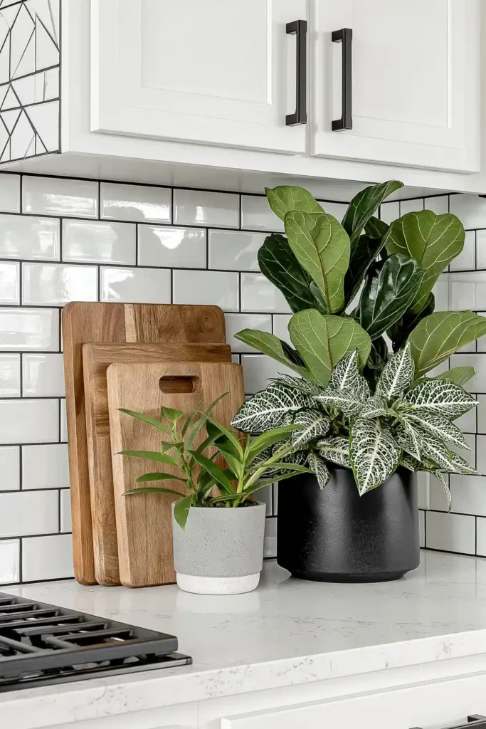 Layered wooden cutting boards leaning against a white subway tile backsplash with small plants on a marble-look kitchen counter.