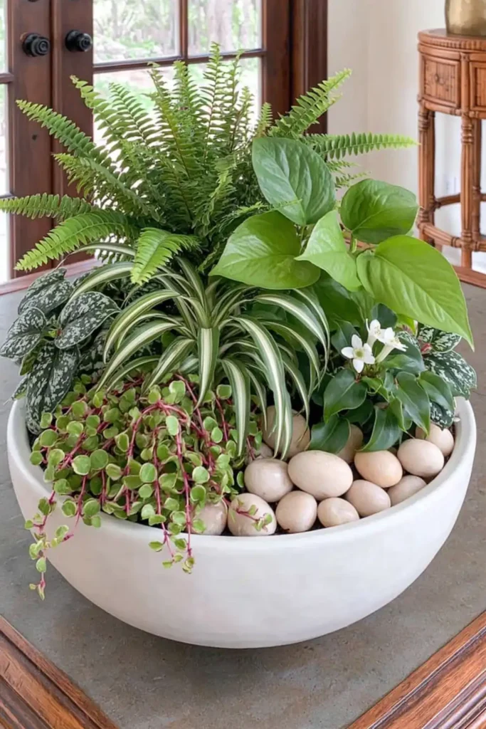 White round bowl planter filled with mixed greenery, fern-like leaves, trailing stems, small white flowers, and pale pebbles.
