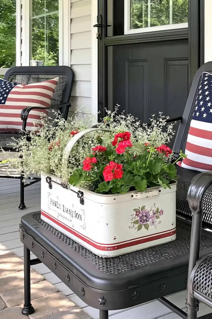 Vintage tin picnic basket planter with red flowers between black porch chairs and patriotic pillows.