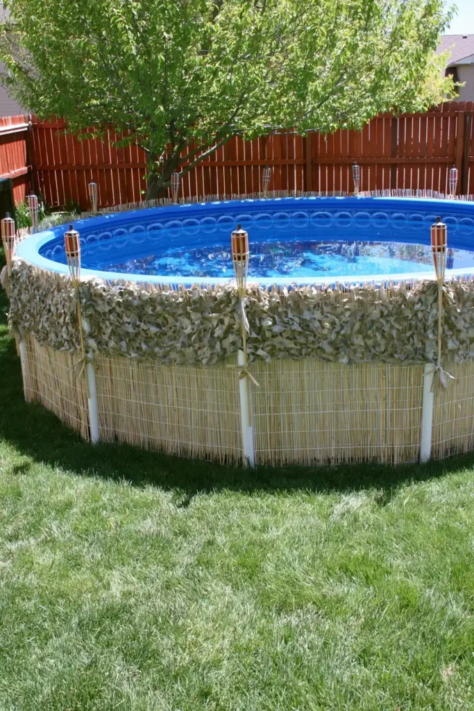 Above-ground pool camouflaged with reed screens, duck blind covering, and citronella torches in a rustic backyard.
