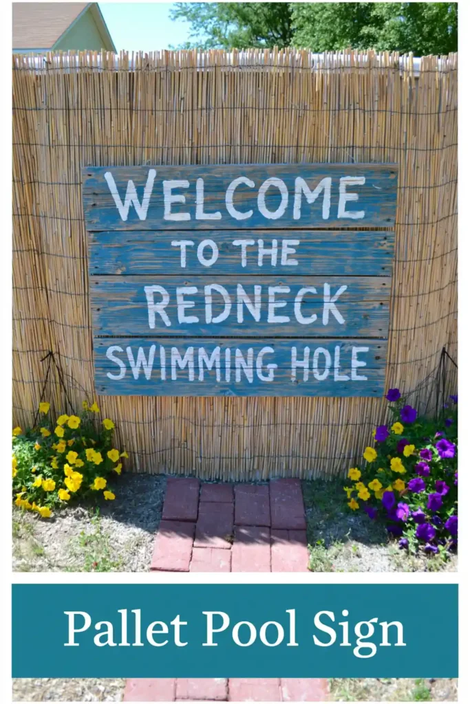 Blue pallet wood welcome sign hanging on reed fencing around an above-ground pool with flowers and a brick path.