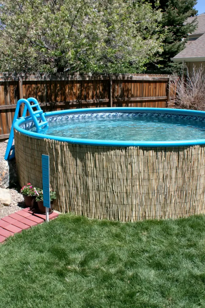 Above-ground pool wrapped with tan reed fencing, blue ladder, pallet sign, potted flowers, and backyard landscaping.