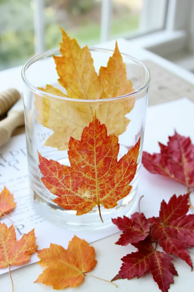 Decoupaged fall leaf candle holder craft with orange and red leaves on a white work surface.
