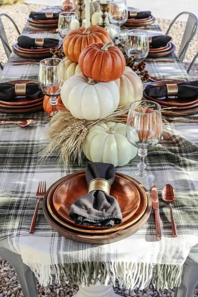 31 Fall Table Decor Ideas for Entertaining in a Warm, Seasonal Style 23 Outdoor orange and gray fall table with plaid fabric, stacked pumpkins, dried wheat, copper cutlery, and dark napkins.