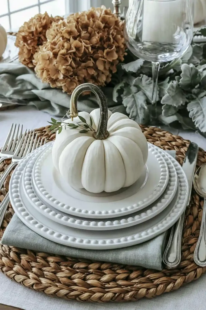 31 Fall Table Decor Ideas for Entertaining in a Warm, Seasonal Style 2 Small white pumpkin centered on layered white plates over a woven charger with a sage napkin and silver flatware.