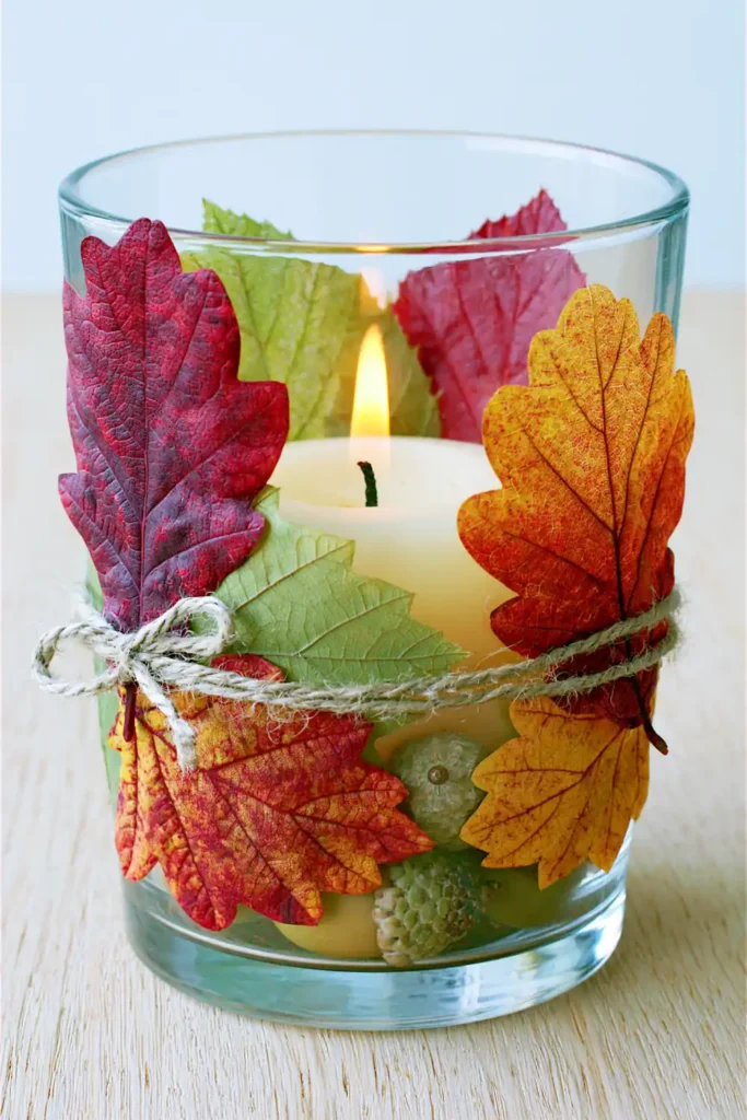 Clear glass candle holder decorated with fall leaves, twine, acorns, and a glowing white candle.