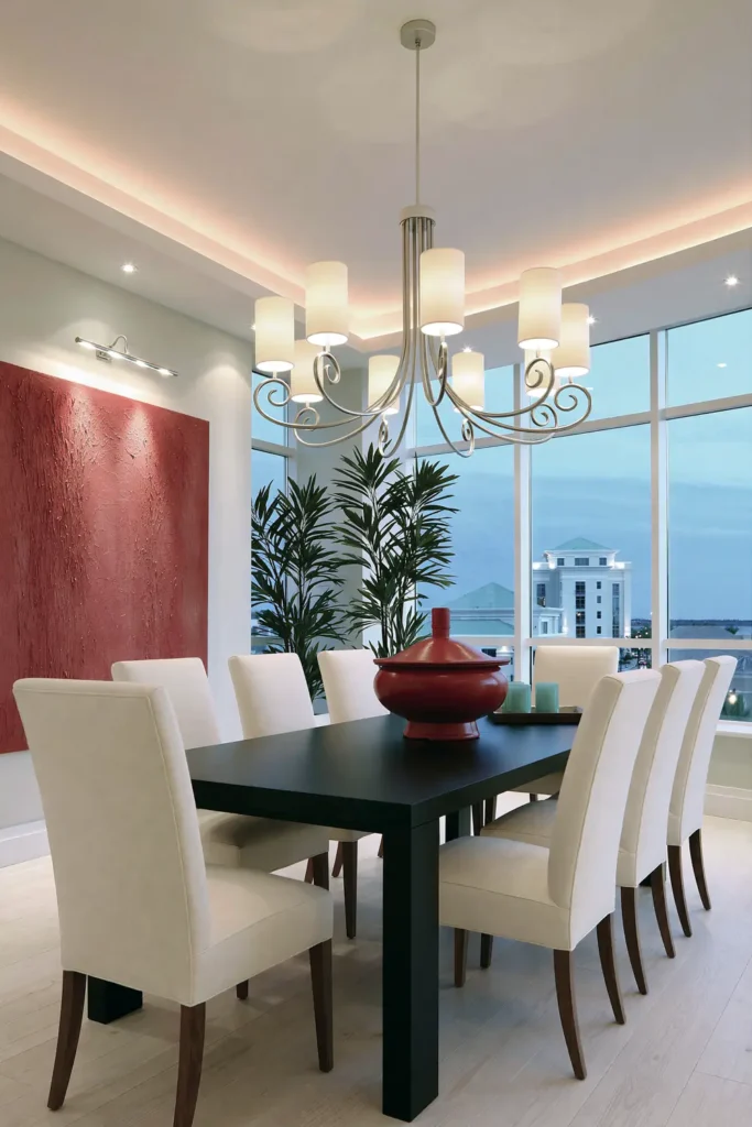 Dining Room Lighting Ideas to Create the Perfect Mood 11 Formal dining room with accent lighting on textured red artwork, a cream-shaded chandelier, black table, and upholstered chairs.