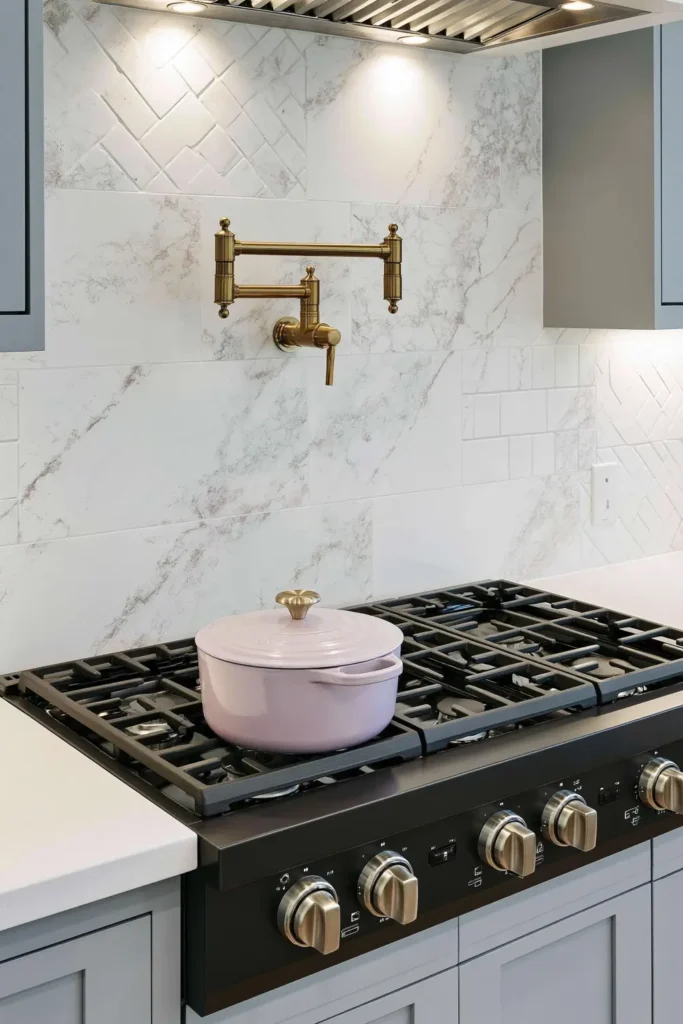 Brass wall-mounted pot filler above a black gas cooktop with a pale veined slab backsplash.