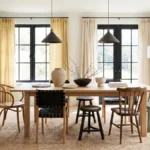 Bright dining room with a light-wood table, mismatched chairs, sculptural tabletop decor, black-framed windows, and natural daylight.