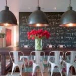Industrial dining room with three oversized dark grey pendant lights above a wooden table, white metal chairs, red flowers, and a chalkboard wall.