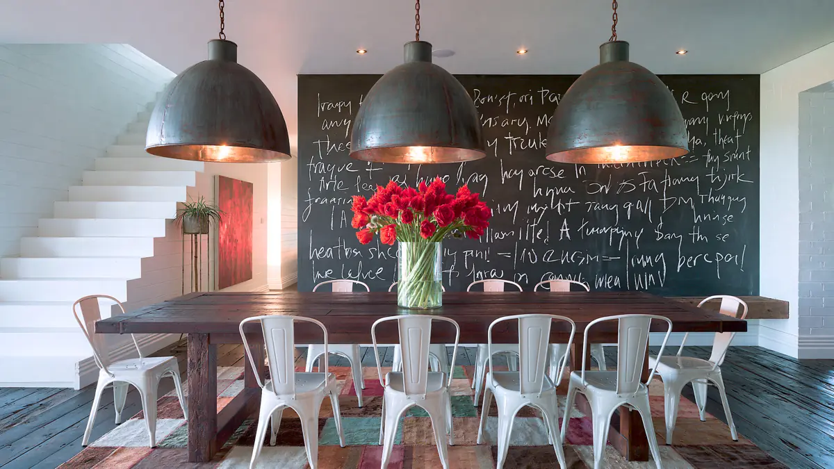 Dining Room Lighting Ideas to Create the Perfect Mood 1 Industrial dining room with three oversized dark grey pendant lights above a wooden table, white metal chairs, red flowers, and a chalkboard wall.