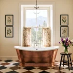 Modern farmhouse bathroom with a copper freestanding tub, diamond-checkered floor, floral café curtains, botanical artwork, and a mountain view.