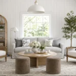 Bright modern farmhouse living room with a gray sofa, white armchairs, rattan shelving, pale wood coffee table, indoor tree, and white vertical wall cladding.