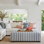 Bright open concept living room with a cream sectional, striped ottoman, white kitchen, exposed ceiling rafters, and an open door facing green foliage.
