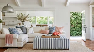 Bright open concept living room with a cream sectional, striped ottoman, white kitchen, exposed ceiling rafters, and an open door facing green foliage.