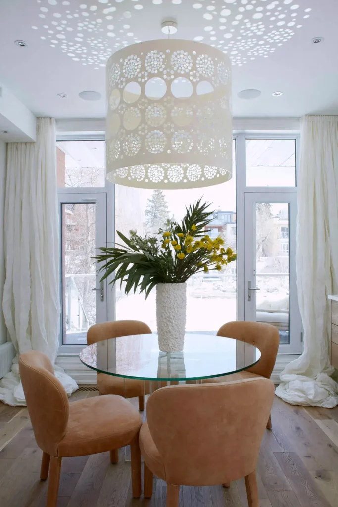 Dining Room Lighting Ideas to Create the Perfect Mood 7 Dining room with a perforated white pendant casting dotted light across the ceiling above a glass table and tan chairs.