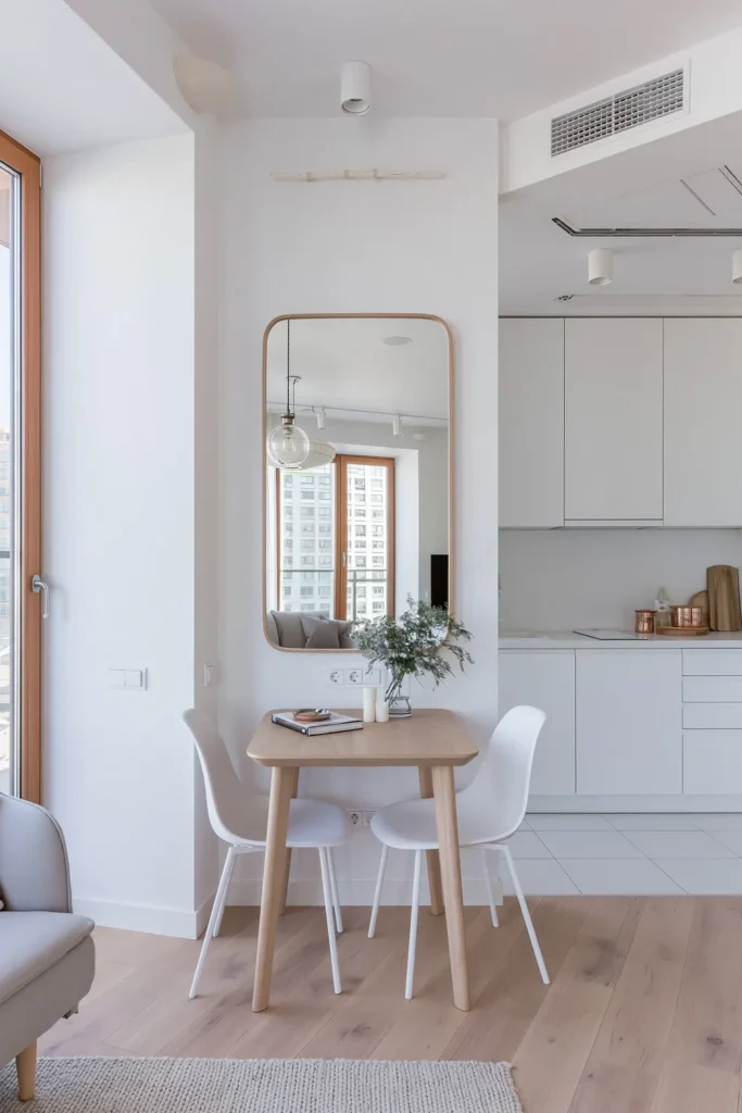 Small Dining Room Ideas for Apartments That Make Every Corner Count 6 Small apartment dining area with a light wood table, white chairs, large wall mirror, pale wood floors, and bright neutral decor.
