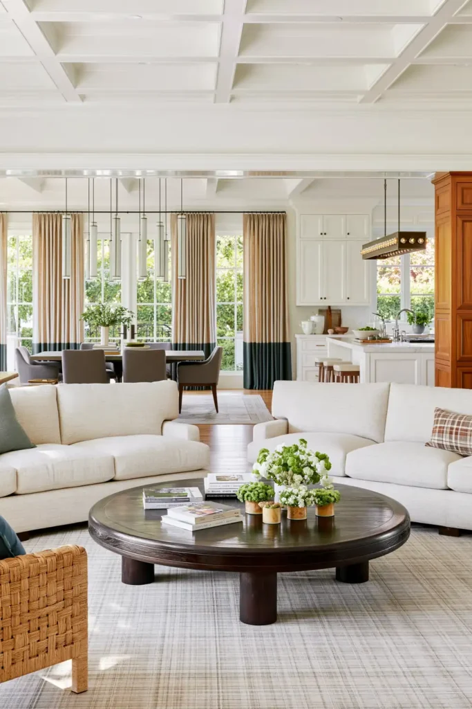 Open living, dining, and kitchen area with cylindrical dining pendants, a rectangular kitchen fixture, beige sofas, and white ceiling beams.