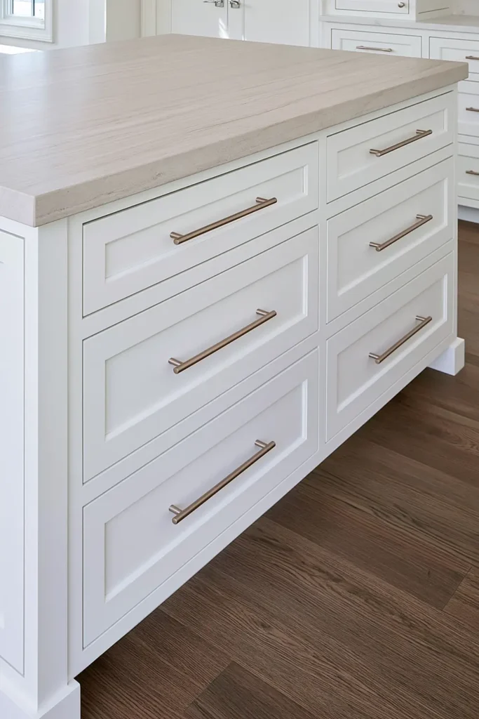 9 Blue and White Kitchen Remodel Ideas with Cozy Gold Accents 3 White shaker kitchen cabinetry with brushed-gold bar pulls, pale stone countertop, and dark stained wood flooring.