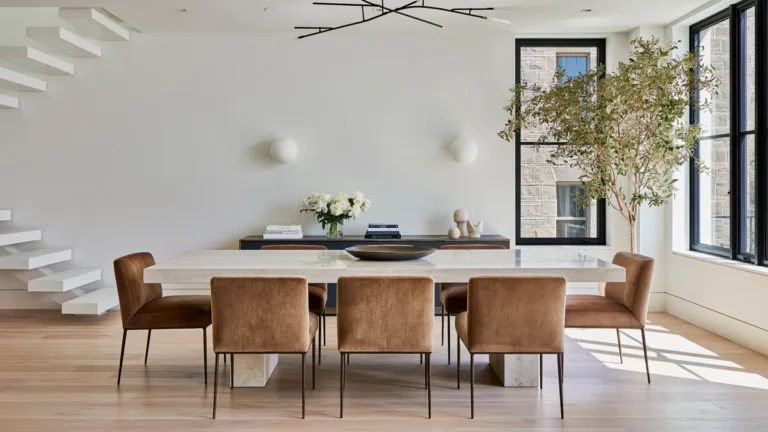8 Formal Dining Room Ideas That Actually Feel Welcoming 2 Welcoming modern formal dining room with a pale stone table, caramel upholstered chairs, floating staircase, dark console, indoor tree, and black-framed windows.