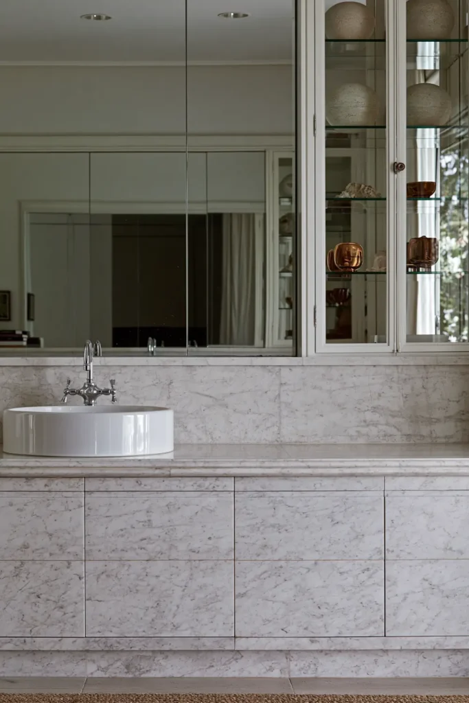 20 Bathroom Shelf Styling Ideas for Beautiful, Practical Storage 8 White marble bathroom with a glass-front display cabinet holding figurines, rounded decorative objects, and amber-toned pieces.