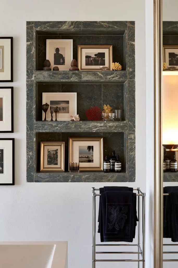 20 Bathroom Shelf Styling Ideas for Beautiful, Practical Storage 4 Blue-gray bathroom storage nook with recessed shelves, framed photographs, small decorative objects, bottles, and dark embroidered towels.