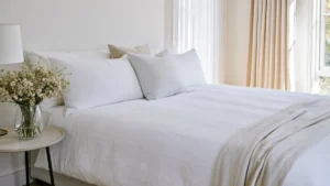 Hotel-style guest bedroom with layered neutral pillows, white bedding, a beige throw, pale flowers, and warm curtains.