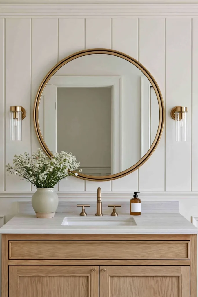 21 Guest Bathroom Decorating Ideas for a Friendly Space 22 Guest bathroom vanity with a large round gold mirror, light wood cabinetry, gold fixtures, and white flowers.
