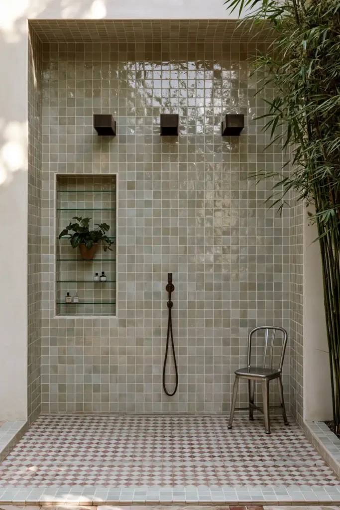 20 Bathroom Shelf Styling Ideas for Beautiful, Practical Storage 7 Mint-tiled shower with a recessed niche, slim glass shelves, small toiletries, leafy plant, silver chair, and checker-pattern floor.