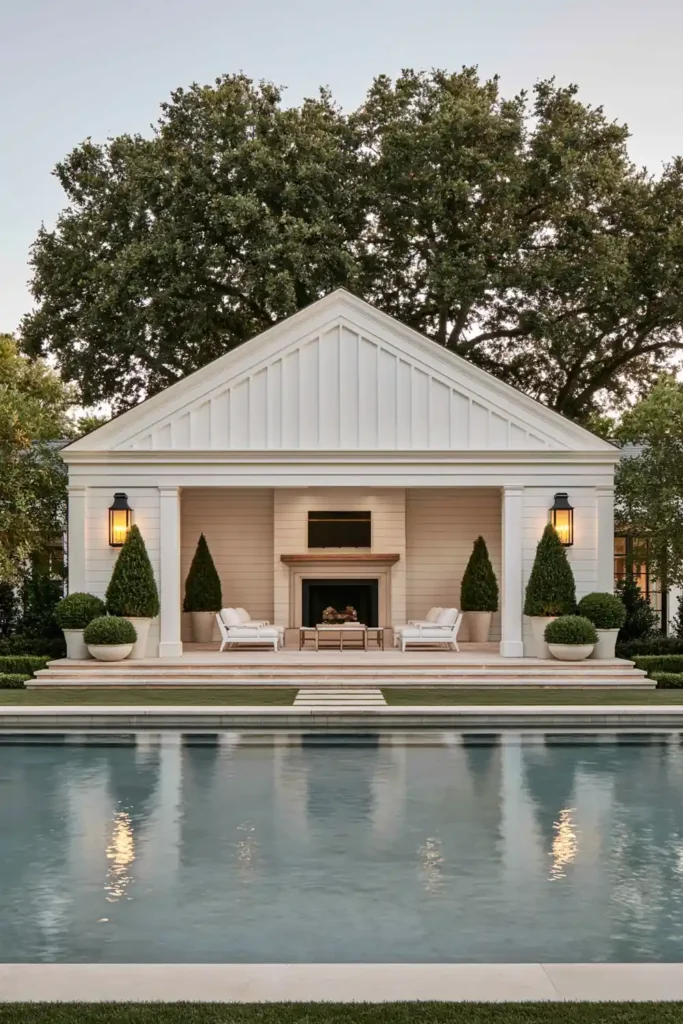 Timeless vs. Trendy Home Decor: 20 Ideas for a Well-Proportioned, Beautiful Home 20 Symmetrical rectangular pool facing a white gabled pool house with a fireplace, lounge chairs, topiary, potted shrubs, and warm wall lanterns.