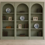Arched built-in bookcase in muted sage with three display bays, books, family photos, and porcelain plates