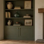 Deep olive alcove shelving beside a wood-burning stove with Shaker cabinet doors, brass knobs, stoneware, books, and botanical art.