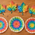 Colorful watercolor coffee filter leaves, three decorated round coffee filters, and a clear spray bottle arranged on a warm wood surface.