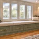 DIY IKEA BESTA window seat with olive-green storage drawers, brass pulls, cream cushions, plantation shutters, patterned pillows, and wall sconces.