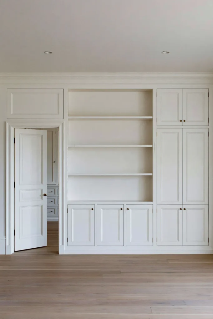 18 Secret Door Ideas That Fit Beautifully Into Your Home 10 White built-in cabinetry with an integrated paneled secret door, open shelving, brass knobs, and pale wood flooring.