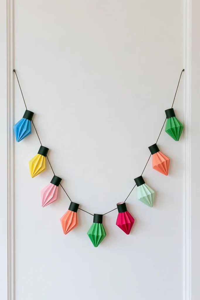 Colorful DIY paper string-light advent calendar decor with folded blue, yellow, pink, orange, green, and magenta paper bulbs hanging along a curved dark cord.