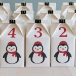 Handmade DIY advent calendar decor featuring white numbered gift boxes with penguin decorations, red hats, scarves, and black binder clips arranged on a wooden table.