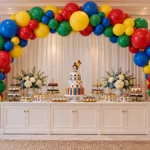 Colorful DIY balloon arch party decor featuring red, blue, green, and yellow balloon clusters framing a white dessert table with a birthday cake, cupcakes, and floral arrangements.