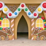 Life-sized gingerbread house entrance with a snowy white roof, red-and-white peppermint columns, colorful gumballs, yellow windows, and oversized gingerbread cookie decorations.