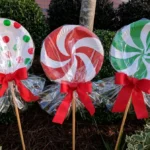 Three DIY giant paper plate lollipops with red, white, and green candy patterns, clear cellophane wrappers, and red ribbon bows displayed among garden shrubs.