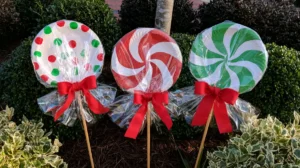 Three DIY giant paper plate lollipops with red, white, and green candy patterns, clear cellophane wrappers, and red ribbon bows displayed among garden shrubs.