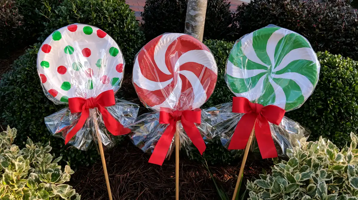 DIY Paper Plate Giant Lollipops Decor: 3 Fun Holiday Display Ideas 1 Three DIY giant paper plate lollipops with red, white, and green candy patterns, clear cellophane wrappers, and red ribbon bows displayed among garden shrubs.