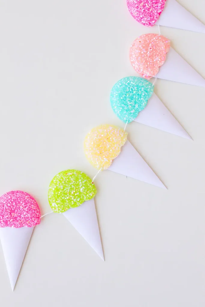 Handmade DIY snow cone garland decor featuring colorful glitter-covered foam snow cones with pointed white paper wrappers arranged on a white tabletop.