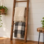 DIY wooden blanket ladder displaying cream and gray buffalo-check blankets against a white paneled wall, styled with hanging greenery and a live-edge wooden side table.