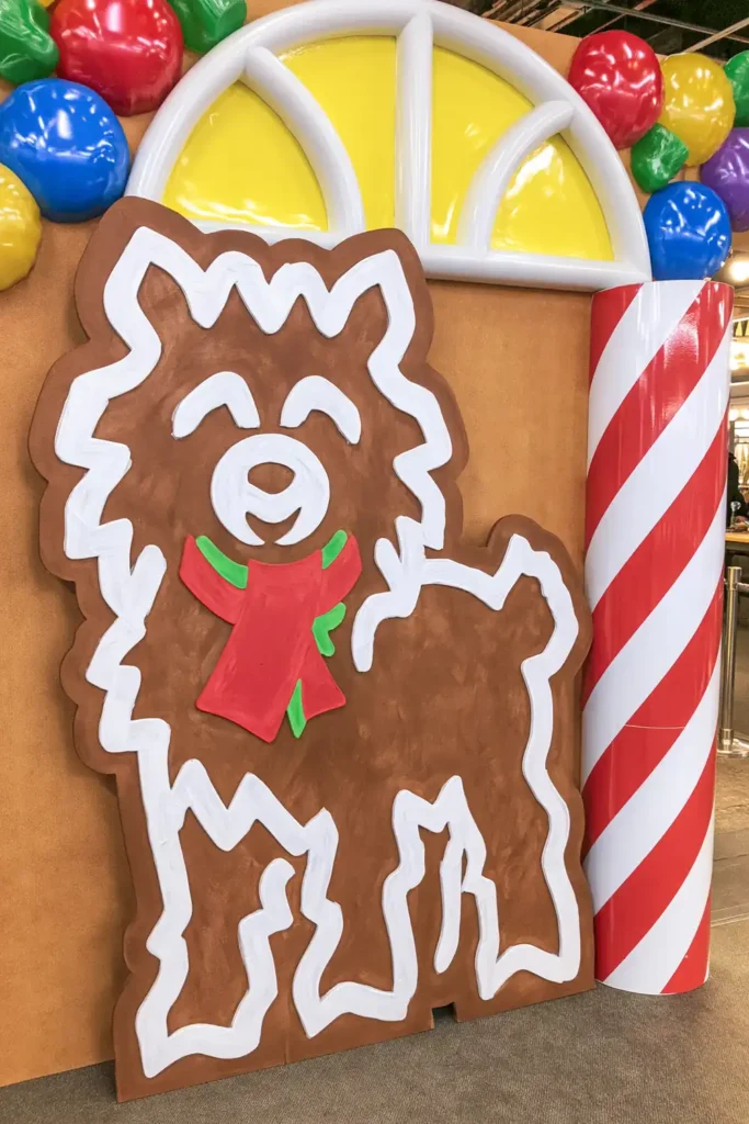 Giant brown animal-shaped gingerbread cookie with white icing outlines and a red-and-green scarf beside a yellow window, colorful gumballs, and peppermint column.