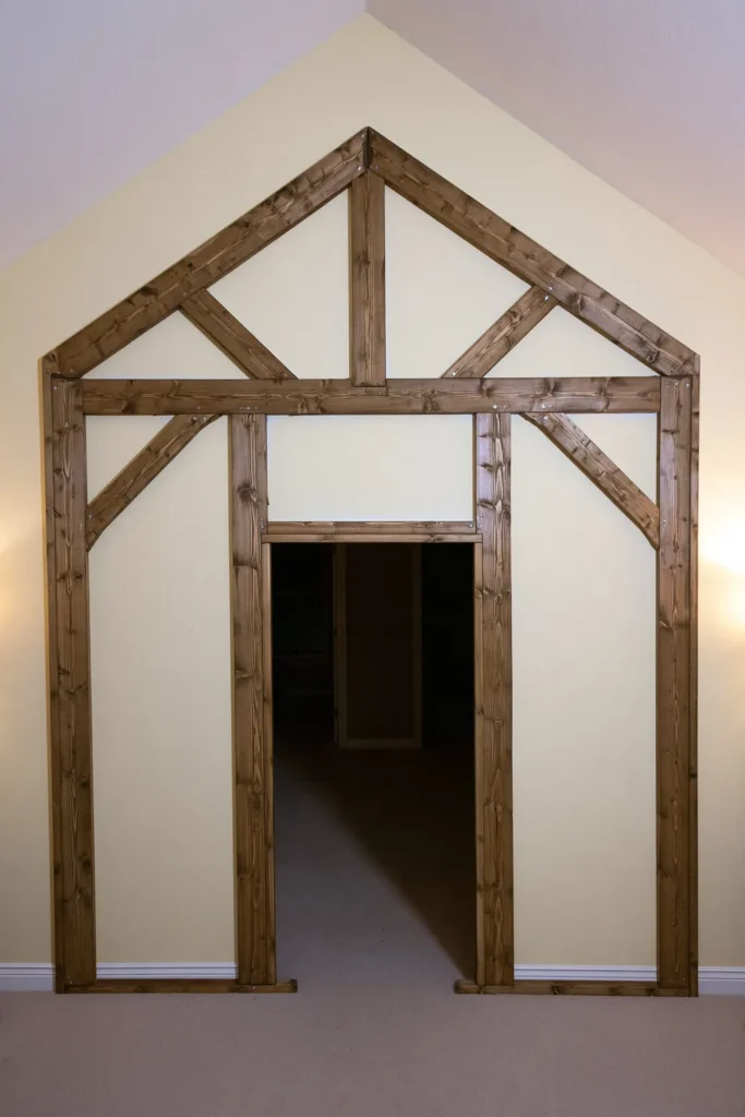 Unfinished life-sized gingerbread house entrance framework built from wooden posts and beams with a pitched roof shape and triangular braces.