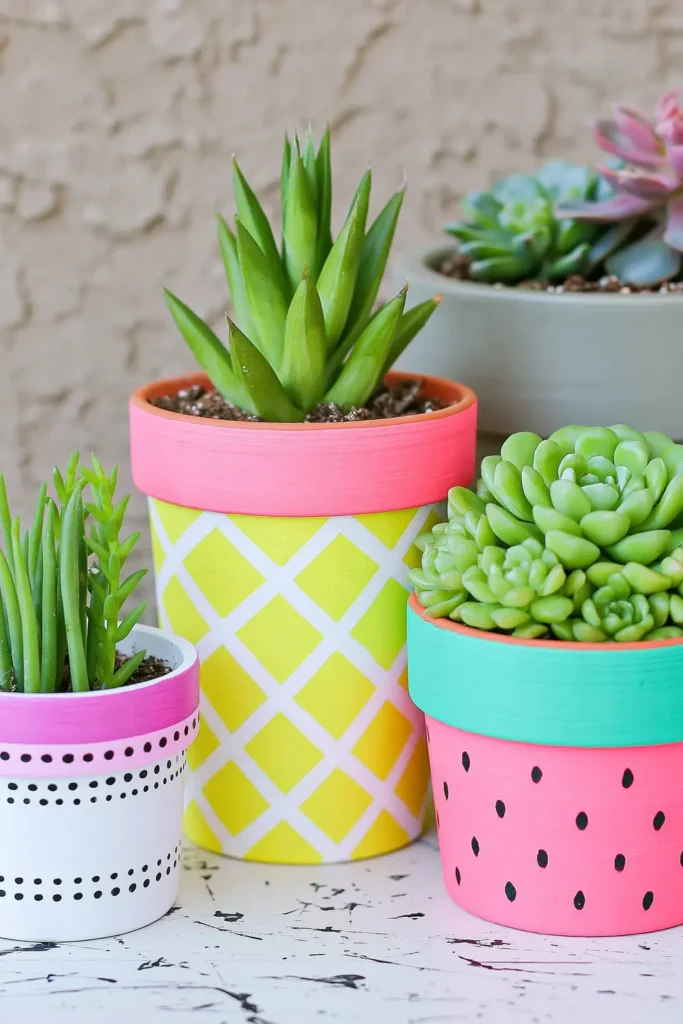 Three colorful summer-painted terracotta planters featuring a yellow pineapple design, a white and magenta fruit design, and a pink watermelon pot filled with green succulents.